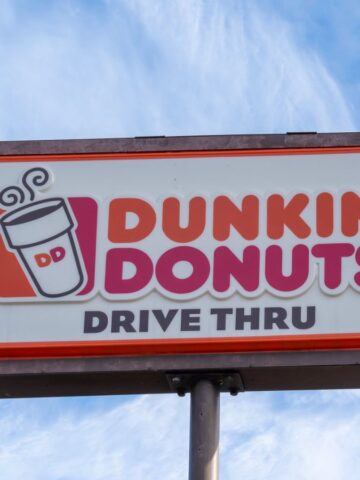 A rectangular Dunkin' Donuts drive-thru sign with a coffee cup illustration, against a blue sky with wispy clouds. The sign has orange, pink, and black text on a white background.