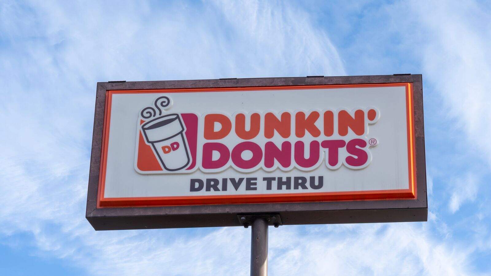 A rectangular Dunkin' Donuts drive-thru sign with a coffee cup illustration, against a blue sky with wispy clouds. The sign has orange, pink, and black text on a white background.