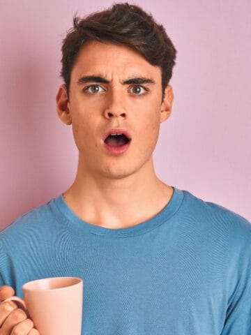 A person with short hair and a surprised expression is holding a pink mug. They are wearing a light blue t-shirt and standing in front of a plain pink background.
