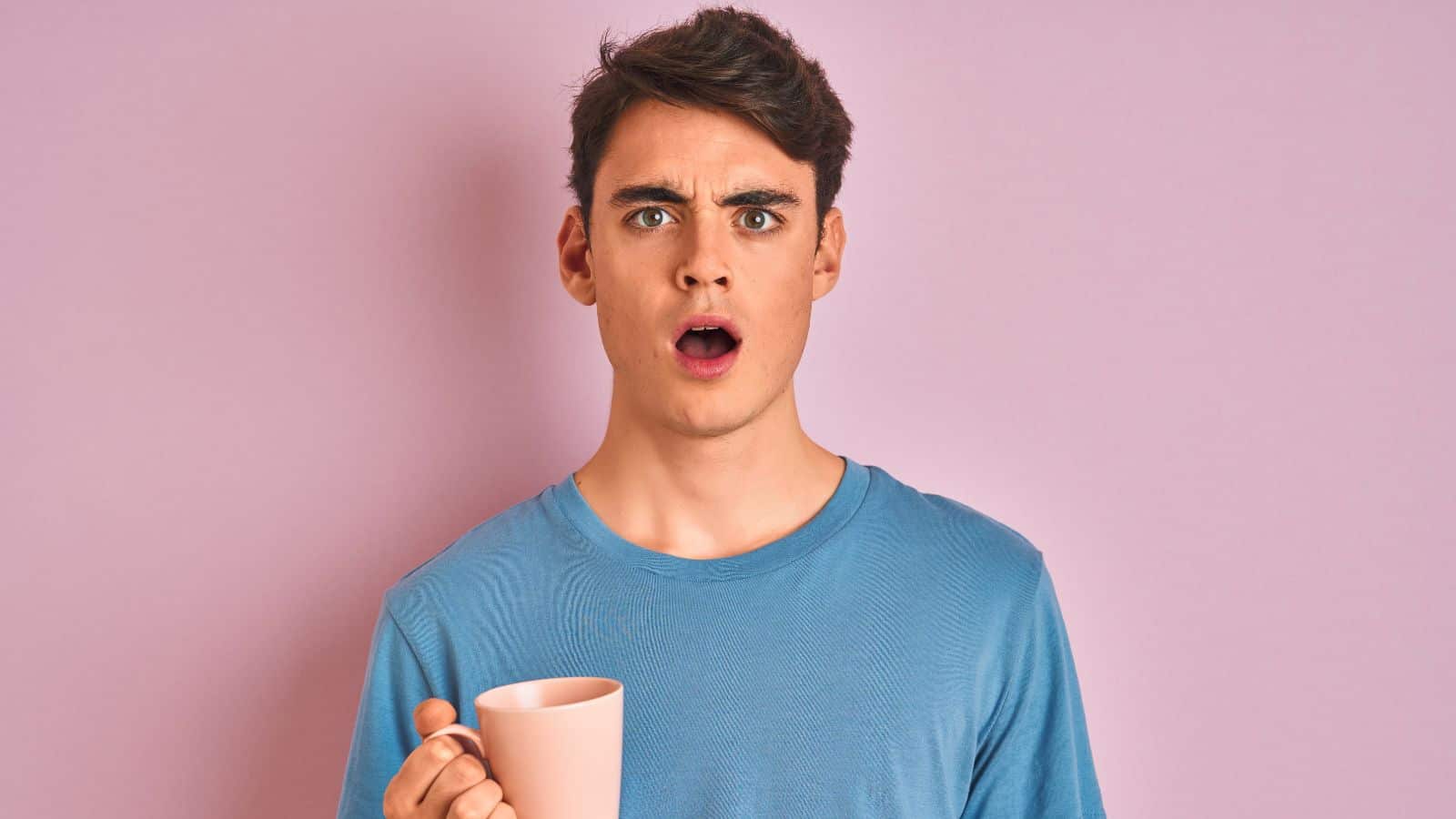 A person with short hair and a surprised expression is holding a pink mug. They are wearing a light blue t-shirt and standing in front of a plain pink background.