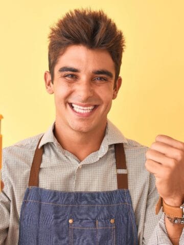 A person wearing a plaid shirt and denim apron smiles while holding a yellow and white reusable coffee cup in one hand and giving a thumbs-up with the other, set against a solid yellow background.