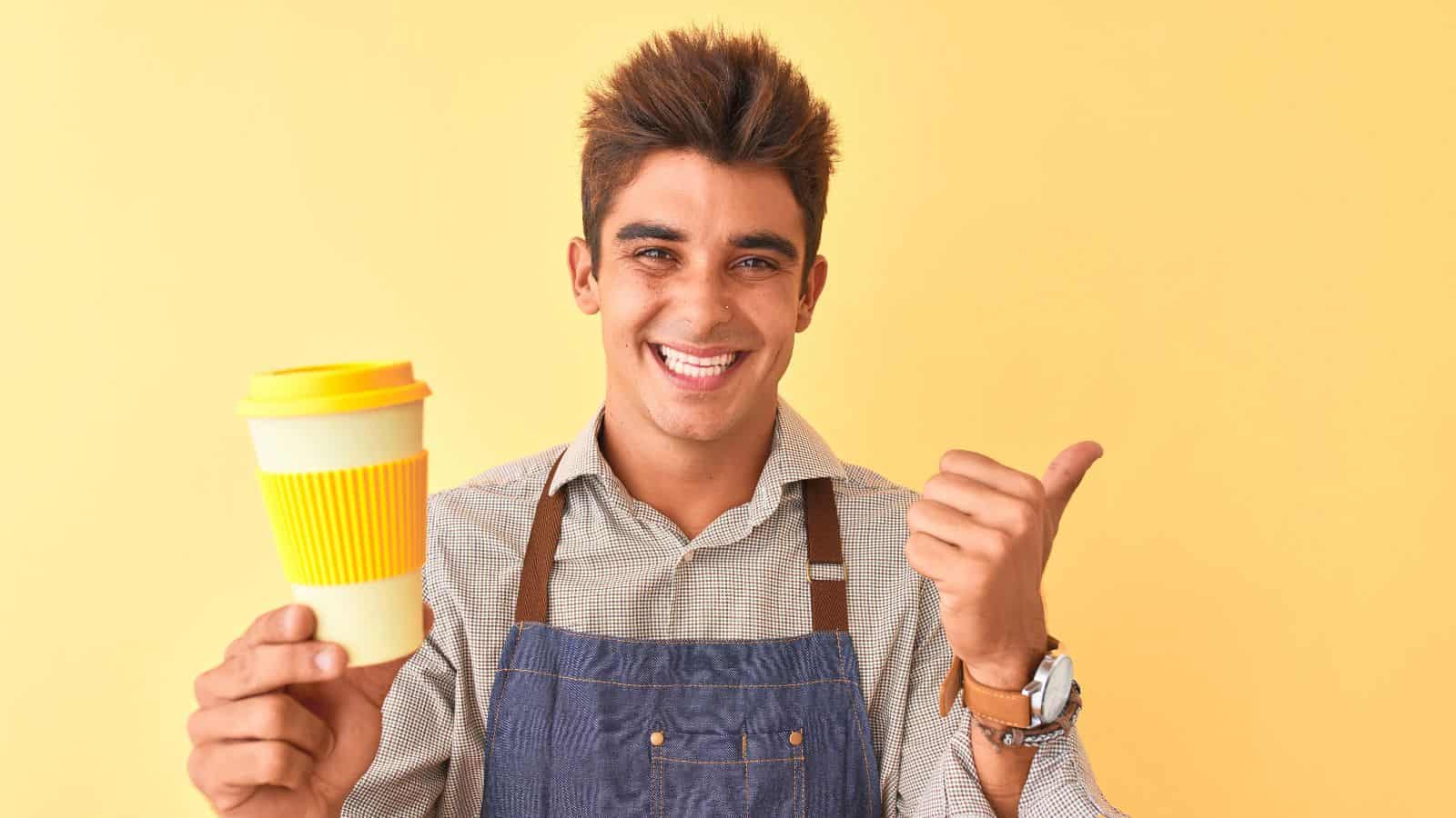 A person wearing a plaid shirt and denim apron smiles while holding a yellow and white reusable coffee cup in one hand and giving a thumbs-up with the other, set against a solid yellow background.