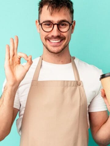 Smiling man in apron holding coffee cup and making an "OK" hand gesture against a teal background, showcasing Tim Hortons essentials.