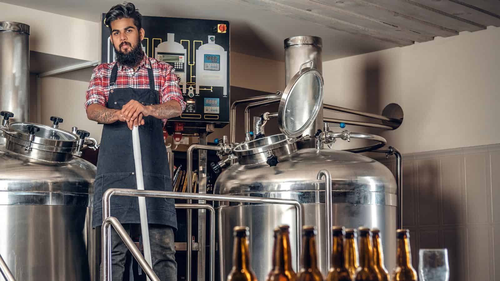 A person with a beard, wearing a red plaid shirt and apron, stands holding a large metal rod in a brewery. Stainless steel brewing tanks and various equipment are in the background. Bottles are in the foreground.