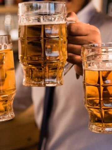 Four people clinking large beer mugs filled with amber-colored beer. They are wearing white shirts and ties, celebrating in what appears to be a bar or pub setting. The focus is on their hands and the mugs.