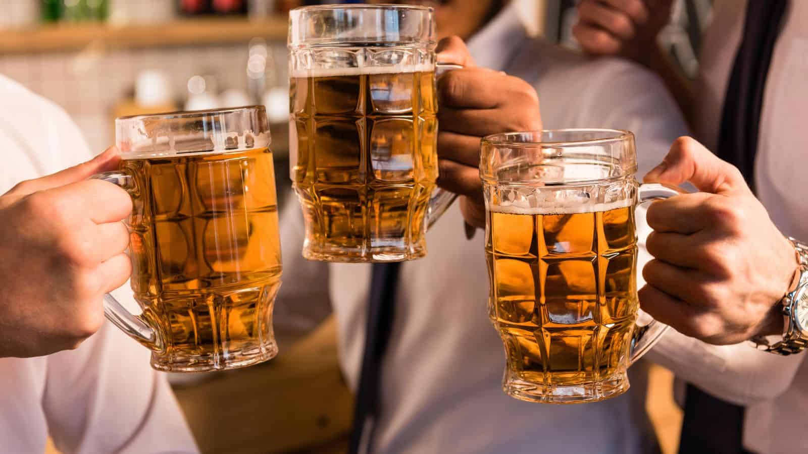 Four people clinking large beer mugs filled with amber-colored beer. They are wearing white shirts and ties, celebrating in what appears to be a bar or pub setting. The focus is on their hands and the mugs.
