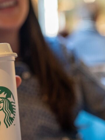 A person is holding a white Starbucks cup with the logo visible. They are seated indoors, with a blurred background showing other people and a warmly lit environment. The person is smiling, and their face is partially out of focus.