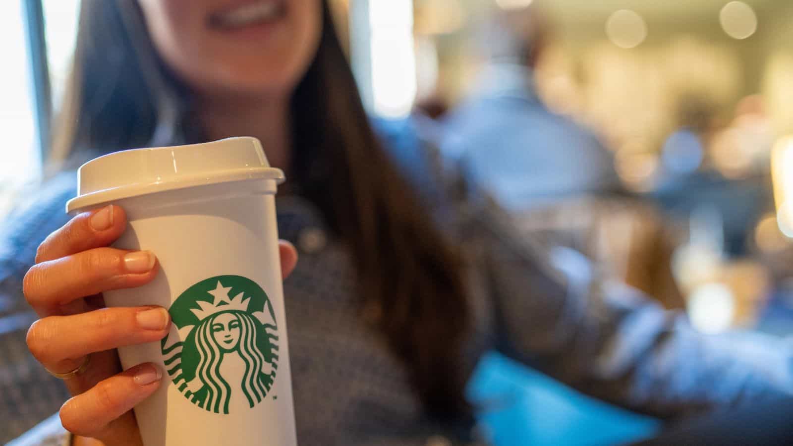 A person is holding a white Starbucks cup with the logo visible. They are seated indoors, with a blurred background showing other people and a warmly lit environment. The person is smiling, and their face is partially out of focus.