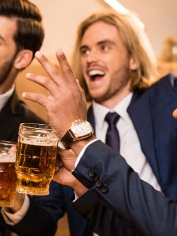 Three men in suits celebrate with beers, holding mugs and smiling. They appear to be in a casual social setting, perhaps at a party or gathering. The mood is lively and cheerful.
