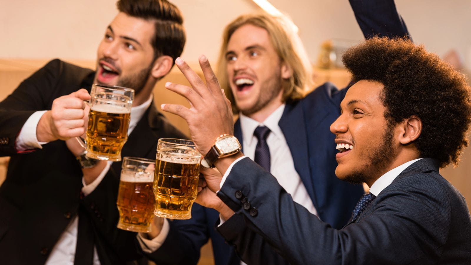 Three men in suits celebrate with beers, holding mugs and smiling. They appear to be in a casual social setting, perhaps at a party or gathering. The mood is lively and cheerful.