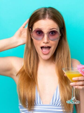 A woman with long hair, wearing sunglasses and a striped top, holds a cocktail in her right hand. The drink has an umbrella garnish. She has an open-mouthed expression. The background is a solid turquoise color.