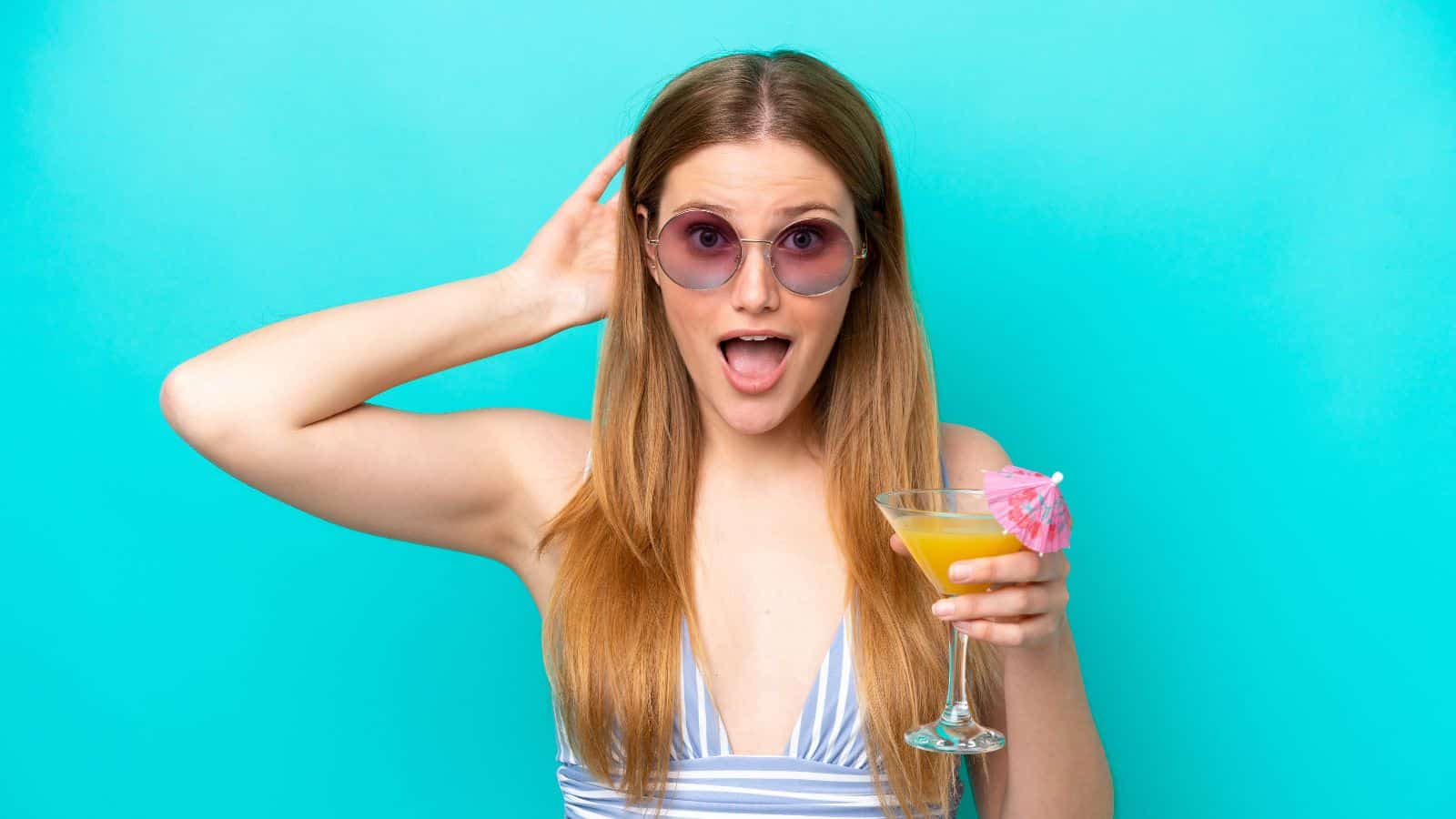 A woman with long hair, wearing sunglasses and a striped top, holds a cocktail in her right hand. The drink has an umbrella garnish. She has an open-mouthed expression. The background is a solid turquoise color.
