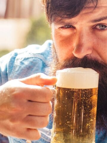 A man with a beard and mustache wears a blue shirt and sits outdoors, holding a glass mug of beer with foam. He looks at the camera while lifting the mug to his mouth.