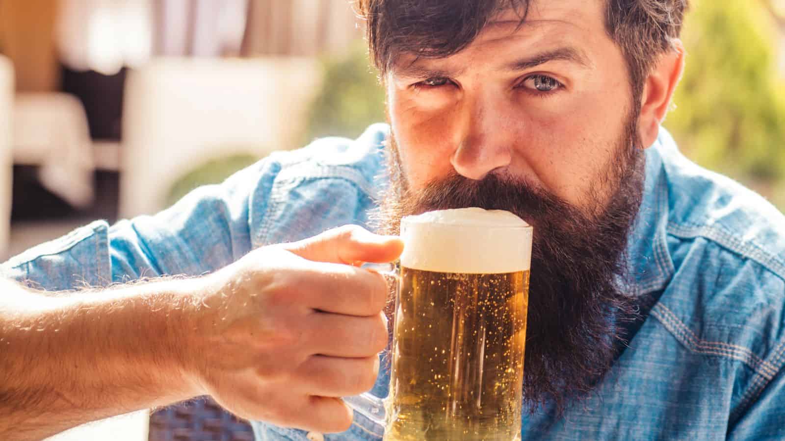 A man with a beard and mustache wears a blue shirt and sits outdoors, holding a glass mug of beer with foam. He looks at the camera while lifting the mug to his mouth.