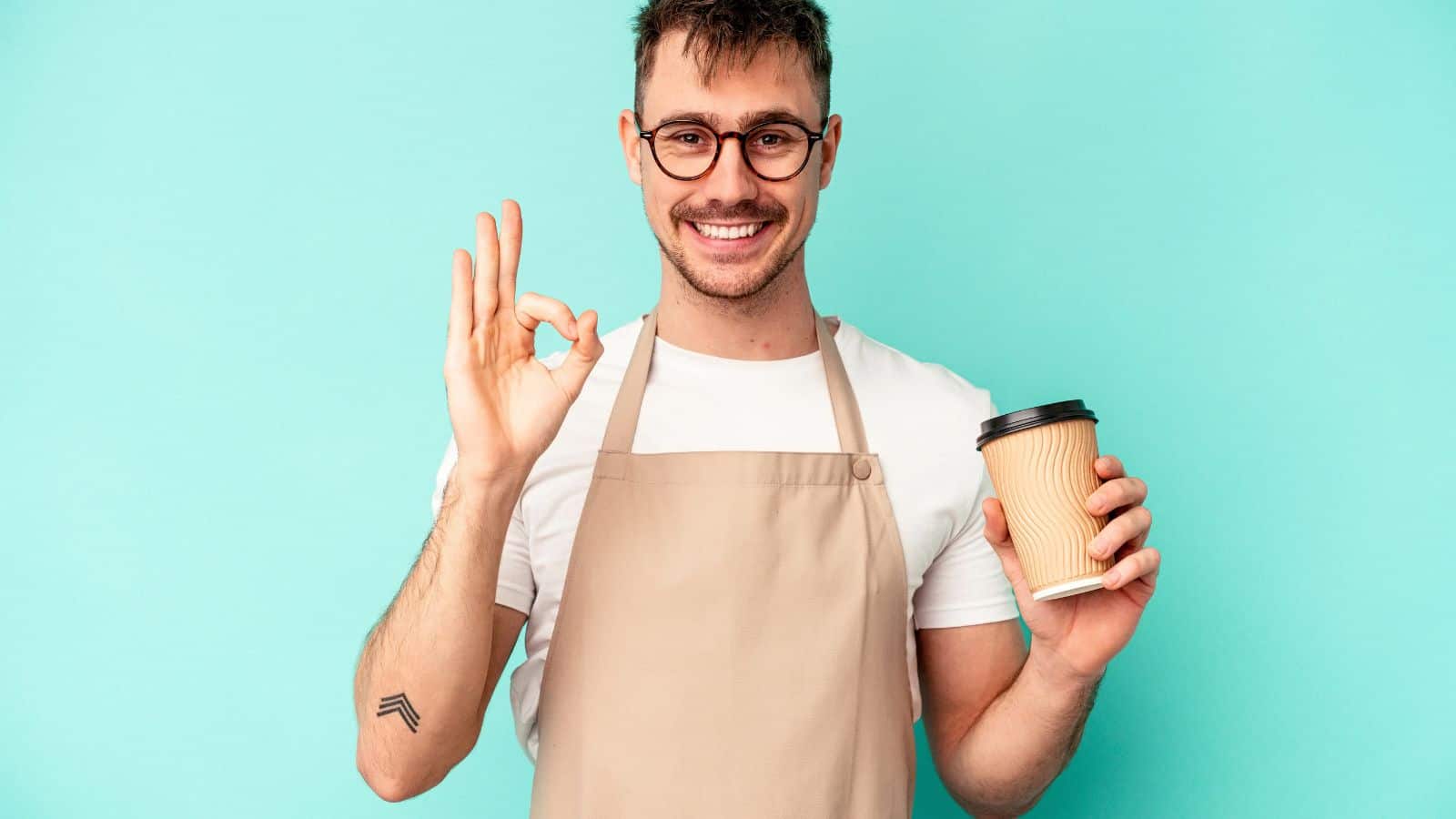 A person with short hair and glasses is wearing a beige apron and holding a takeaway coffee cup. They are making an "OK" gesture with their other hand. The background is a solid light blue color.