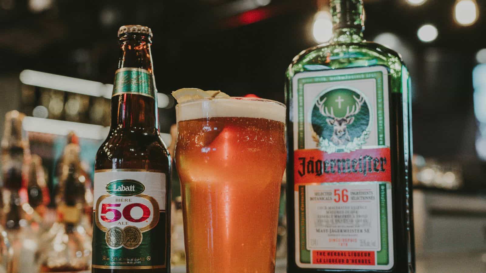 A bottle of Labatt 50 ale and a glass of beer with lemon wedges are on a bar counter, next to a bottle of Jägermeister. The background is blurred with warm, ambient lighting.