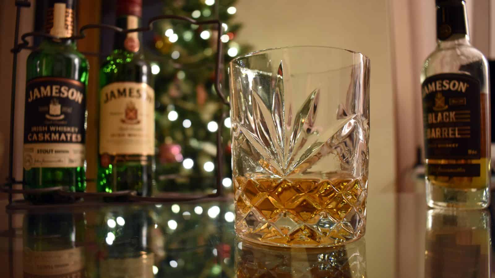A glass with a small amount of whiskey sits on a reflective table. Bottles of Jameson whiskey are in the background, with a decorated, lit Christmas tree slightly out of focus.