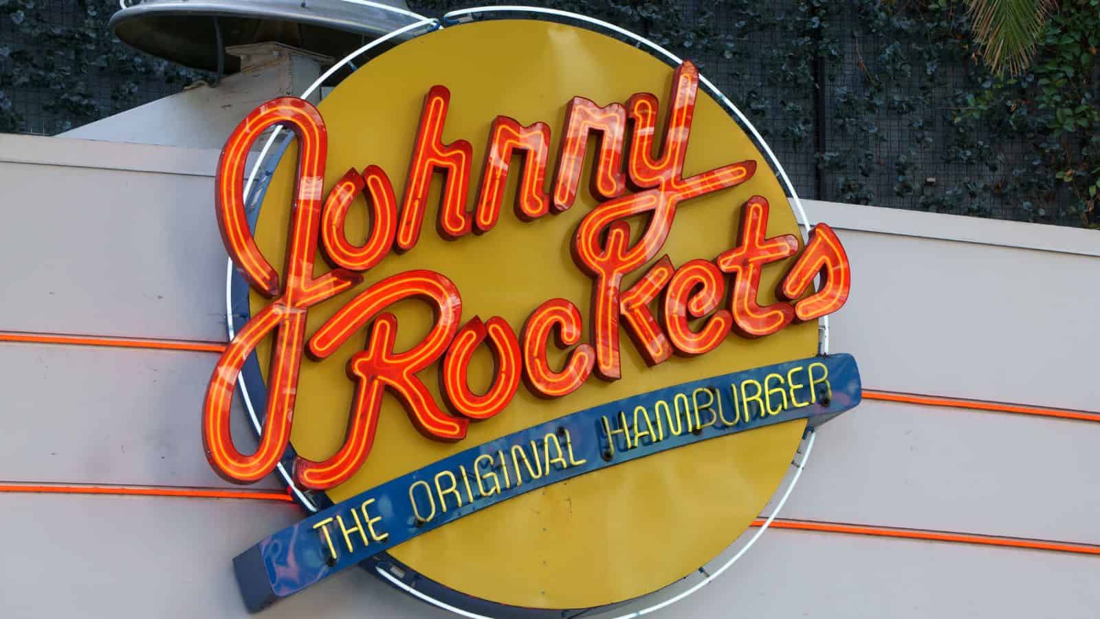 A round yellow and red sign for Johnny Rockets, with the text “Johnny Rockets” in red script and “The Original Hamburger” in blue, mounted on a wall with some orange neon lights.