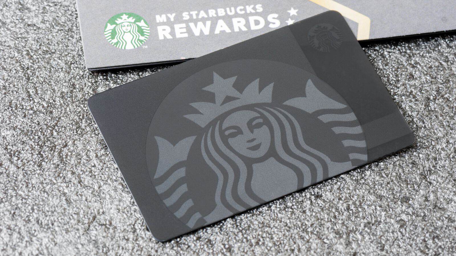 A Starbucks rewards card featuring a stylized mermaid logo is placed on a textured gray surface. Part of a paper with the words &ldquo;My Starbucks Rewards&rdquo; and a partial mermaid logo is visible behind the card.