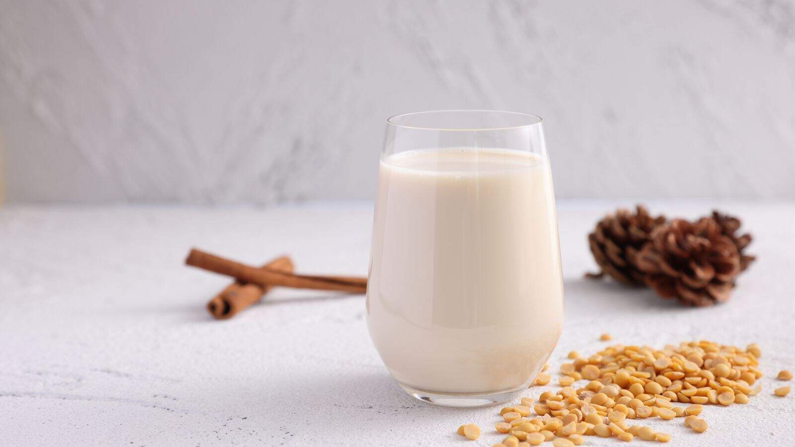 A glass of creamy liquid sits on a white surface next to scattered pine nuts. In the background, there are two cinnamon sticks and a pinecone, all against a light gray textured backdrop.