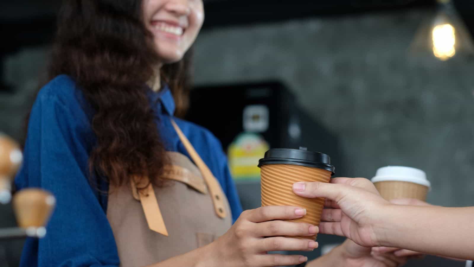 A person wearing a blue shirt and apron hands a brown, ripple-wall coffee cup with a black lid to another individual. The setting appears to be a coffee shop, with blurred background details.