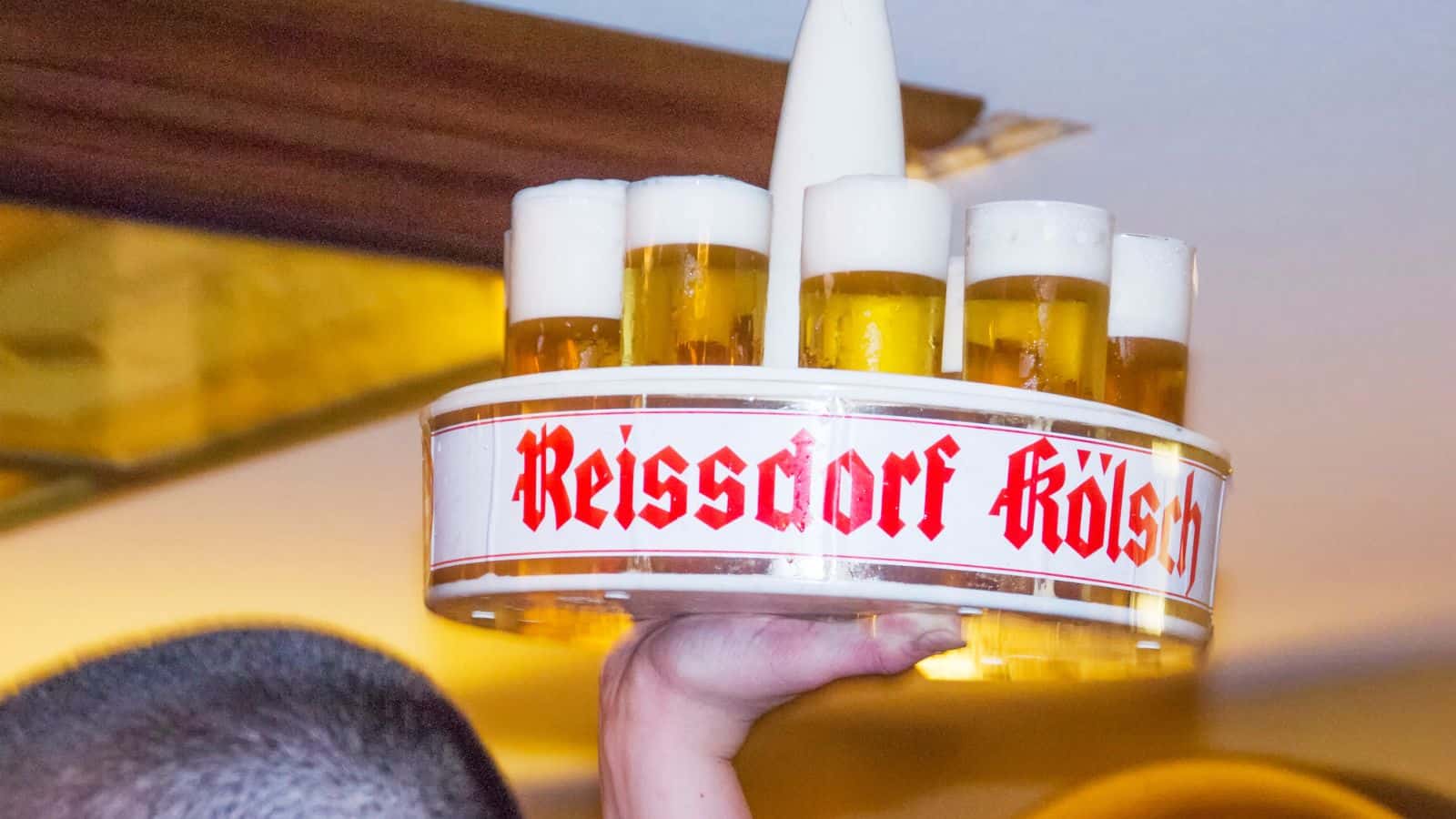 A hand holds up a round tray with eight glasses of beer, each topped with a head of foam. The tray has the words "Reissdorf Kölsch" written in red gothic font, indicating a specific type of beer.