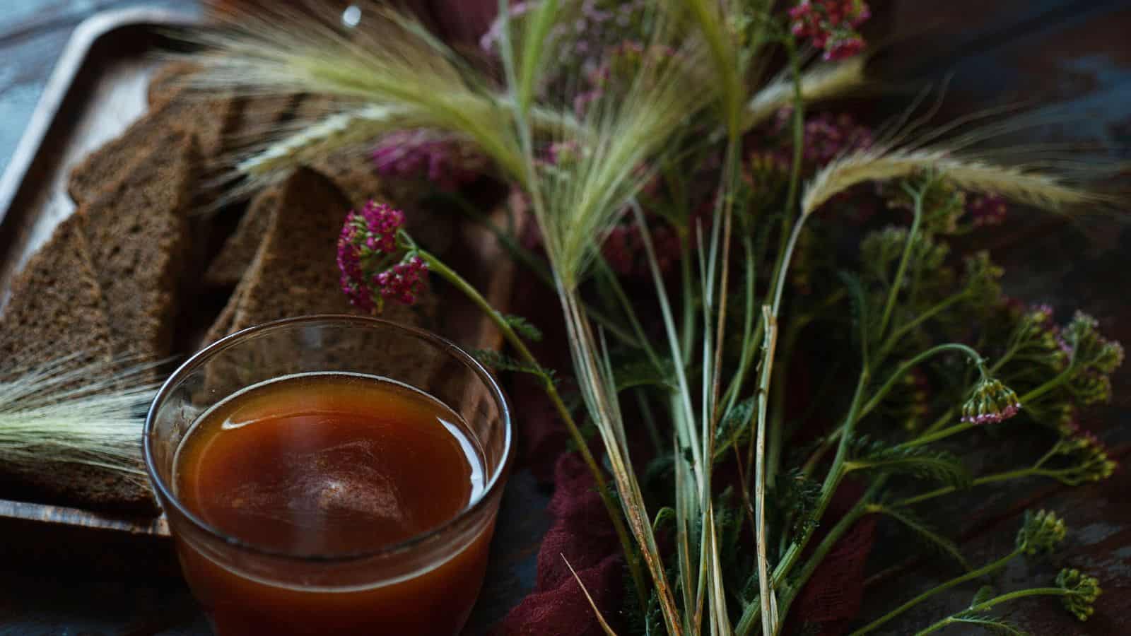 A glass of dark liquid sits on a table next to slices of dark bread and sprigs of wildflowers and grasses. The setting conveys a rustic atmosphere with dim lighting.