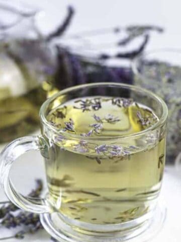 A clear glass cup filled with light yellow herbal tea, garnished with lavender flowers, sits in front of a glass teapot. Dried lavender and a small glass container are nearby on a white surface.