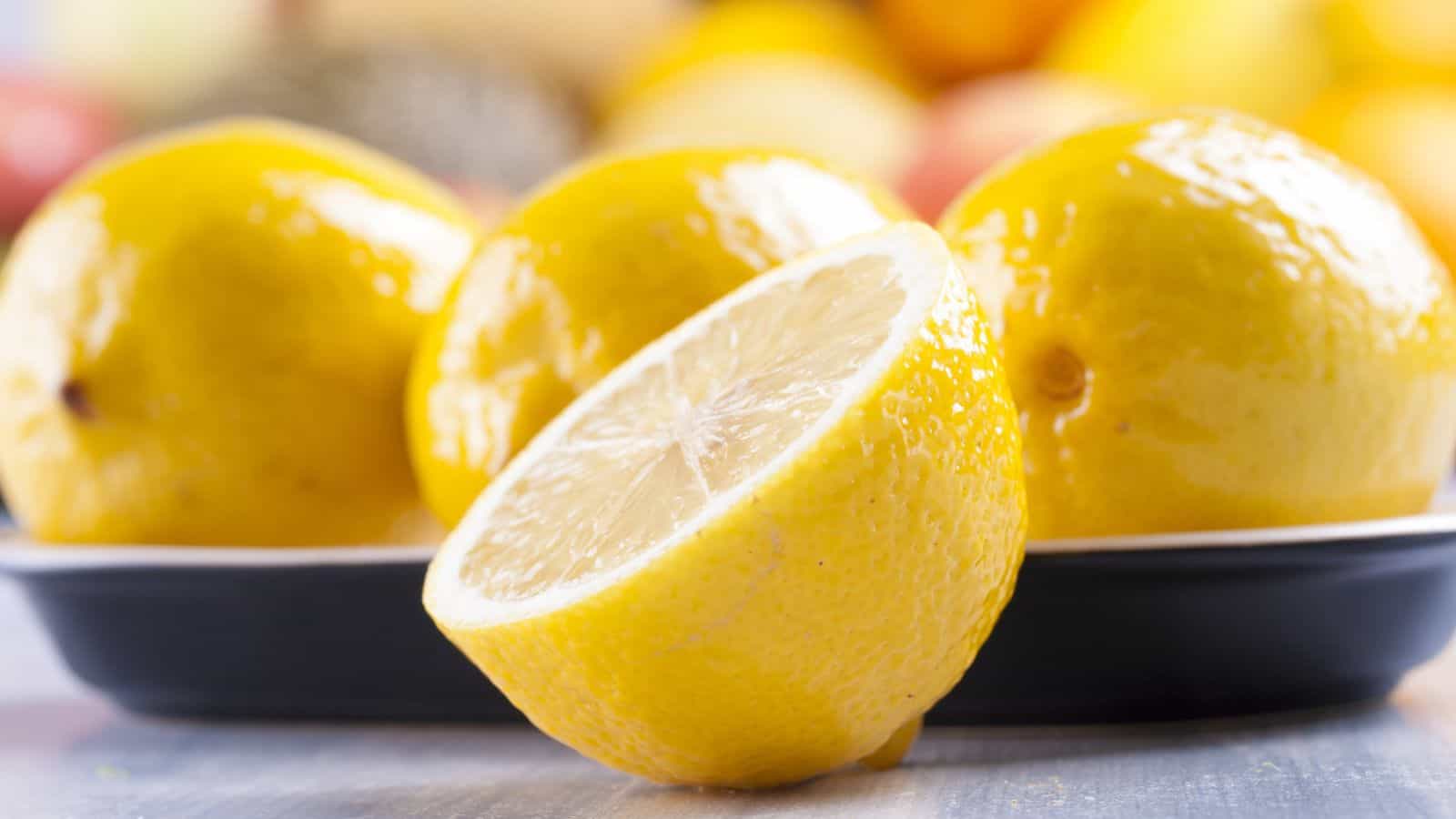 A close-up of three whole lemons and one halved lemon placed on a plate, with a blurred background of more fruits.