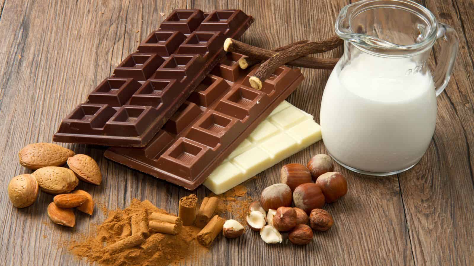A wooden surface with three chocolate bars (dark, milk, and white), a glass jug of milk, almonds, hazelnuts, cinnamon sticks, and a pile of cocoa powder.