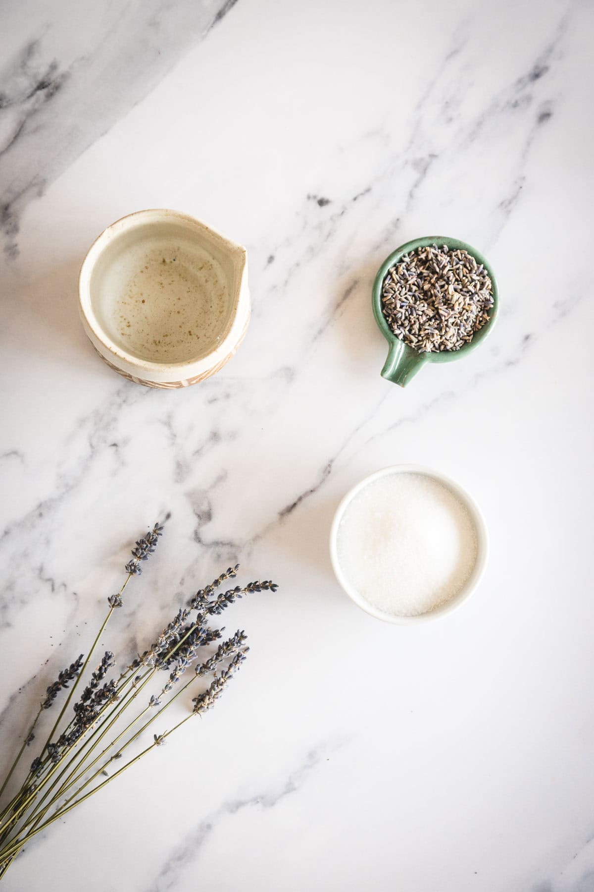 Ingredients needed to make lavender simple syrup.  A ceramic pitcher, a small green cup of dried lavender, a white bowl of granulated sugar, and sprigs of fresh lavender are arranged on a white marble surface.