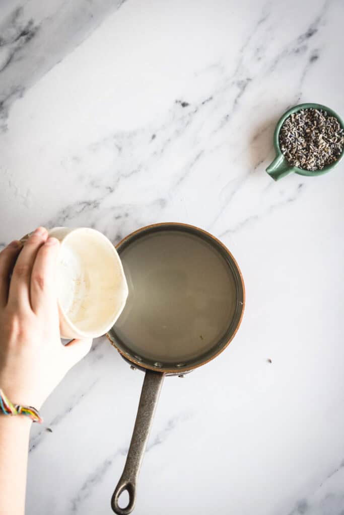 A hand pours a white powder from a small bowl into a saucepan filled with liquid on a marble surface. A green cup containing dried lavender sits nearby. Steps to make the lavender syrup. 
