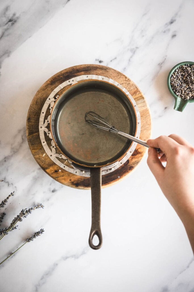 A hand holds a small whisk inside a metal saucepan with lavender simple syrup, placed on a wooden trivet. The scene is set on a marble surface, with dried lavender sprigs and a small green bowl of lavender buds nearby.