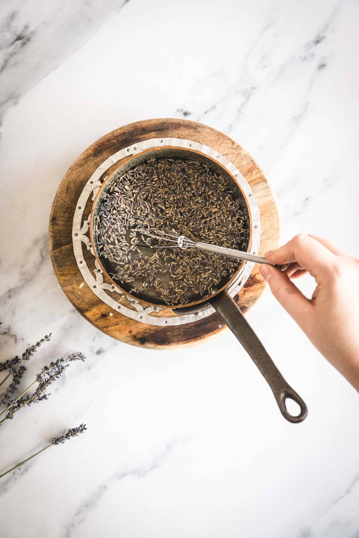A hand stirs dried lavender buds with a spoon in a metal saucepan placed on a wooden trivet for the lavender syrup, with some sprigs of lavender nearby on a white marble surface.