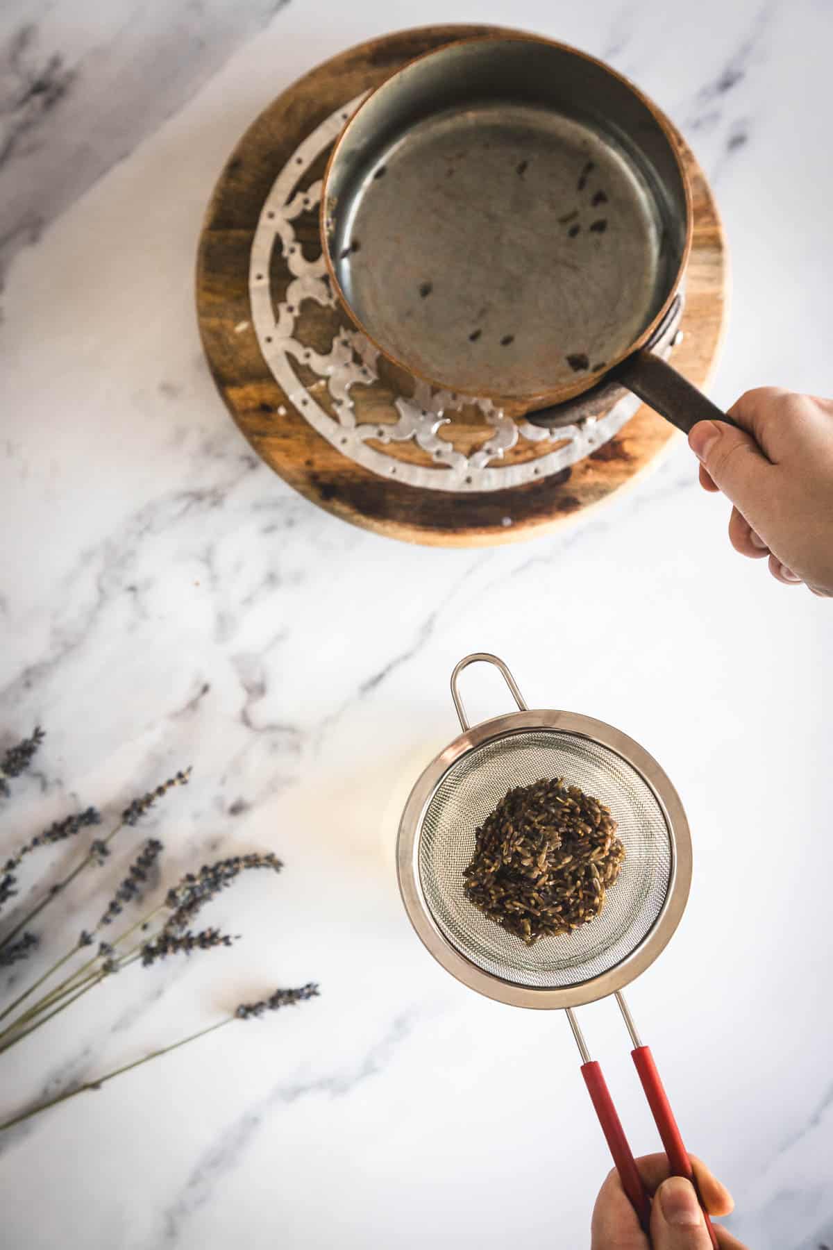 A person holds a metal strainer with dried tea leaves over a pot of hot water placed on a round wooden trivet on a marble countertop; lavender sprigs are nearby.