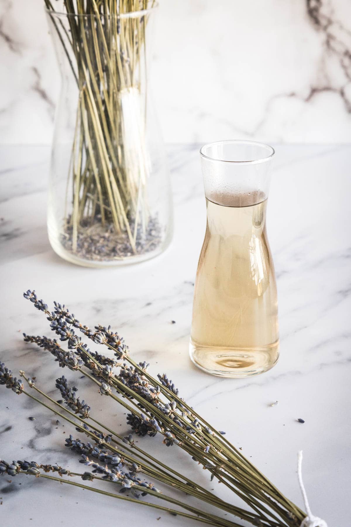 A glass carafe filled with lavender simple syrup on a marble surface beside a bundle of dried lavender stems and a vase containing more dried lavender. Scattered lavender buds are visible on the table.