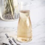 A clear glass carafe filled with light-colored liquid sits on a marble surface, with a bundle of dried lavender and a vase of lavender stems in the background.