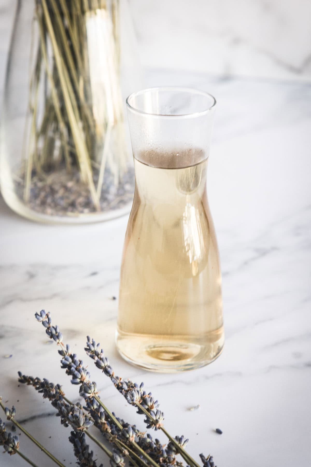 A jar with lavender syrup sits on a white marble surface. Dried lavender sprigs and a jar of dried lavender are nearby in the background.