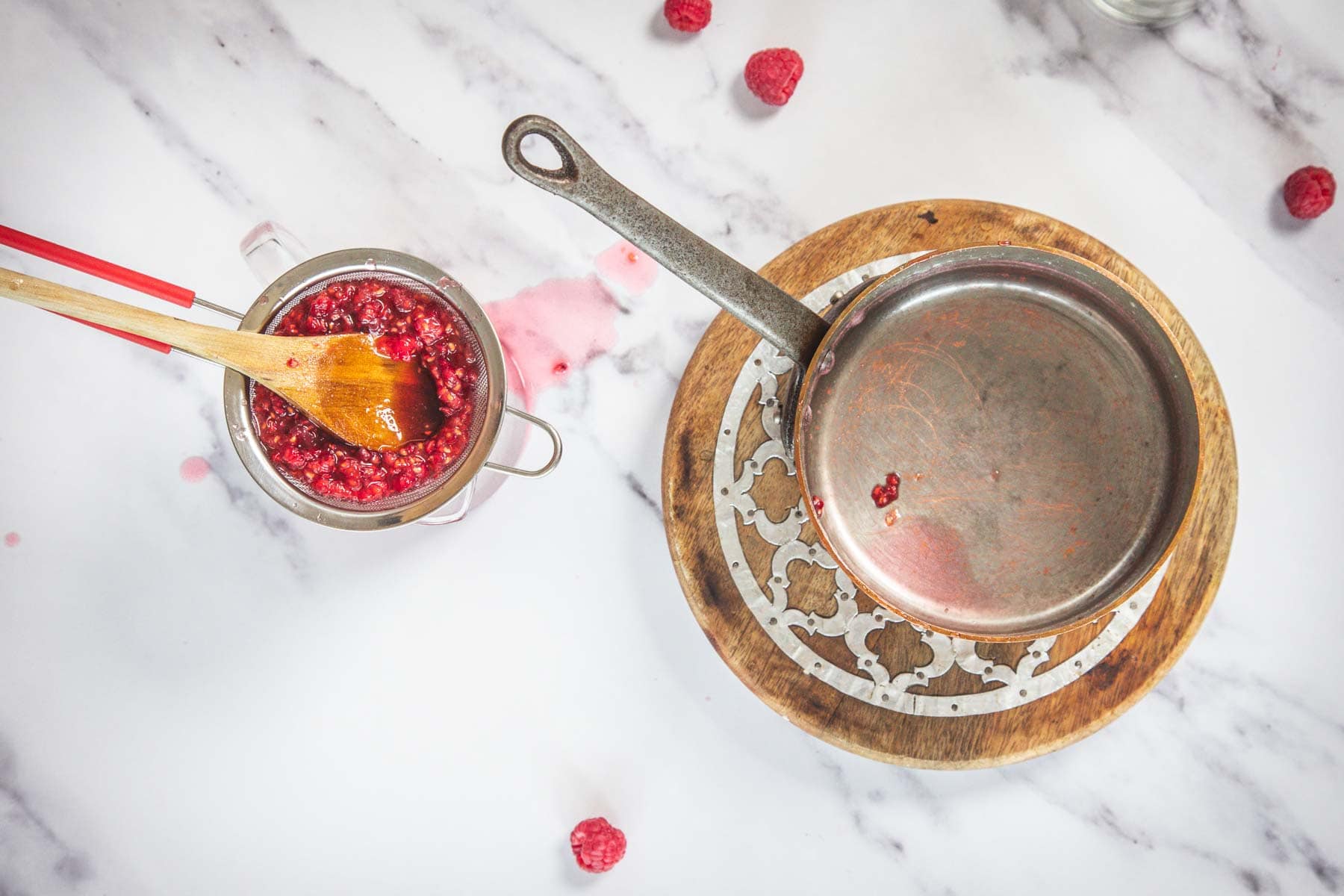 A small saucepan sits on a wooden trivet next to a metal strainer containing mashed raspberries and a wooden spoon. A few raspberries and pink juice are on the white marble countertop.