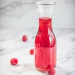 A clear glass carafe filled with red liquid, surrounded by four raspberries on a white marble surface. The background is also a white marble pattern.