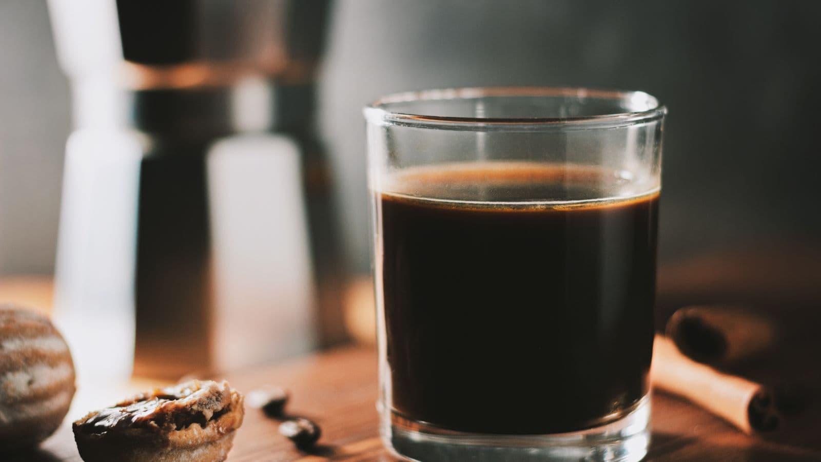 A glass of dark coffee sits on a wooden surface, surrounded by a cinnamon stick, a halved nutmeg, coffee beans, and a blurred moka pot in the background.