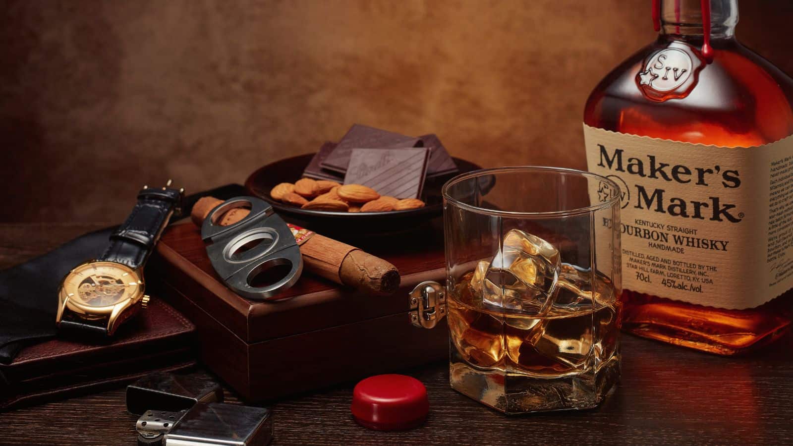 A bottle of Maker’s Mark bourbon whisky, a glass of whisky with ice, a cigar, a cigar cutter, a watch, a lighter, a bowl with almonds and chocolate, and a wallet on a dark wooden surface.