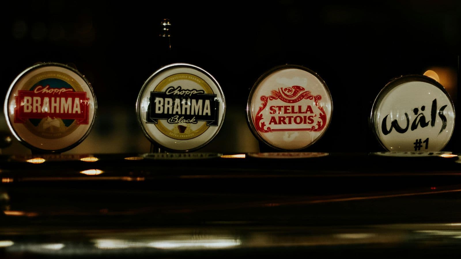 Four beer taps are shown in a dark setting. From left to right, the taps display labels for Chopp Brahma, Chopp Brahma Black, Stella Artois, and Wäls #1.