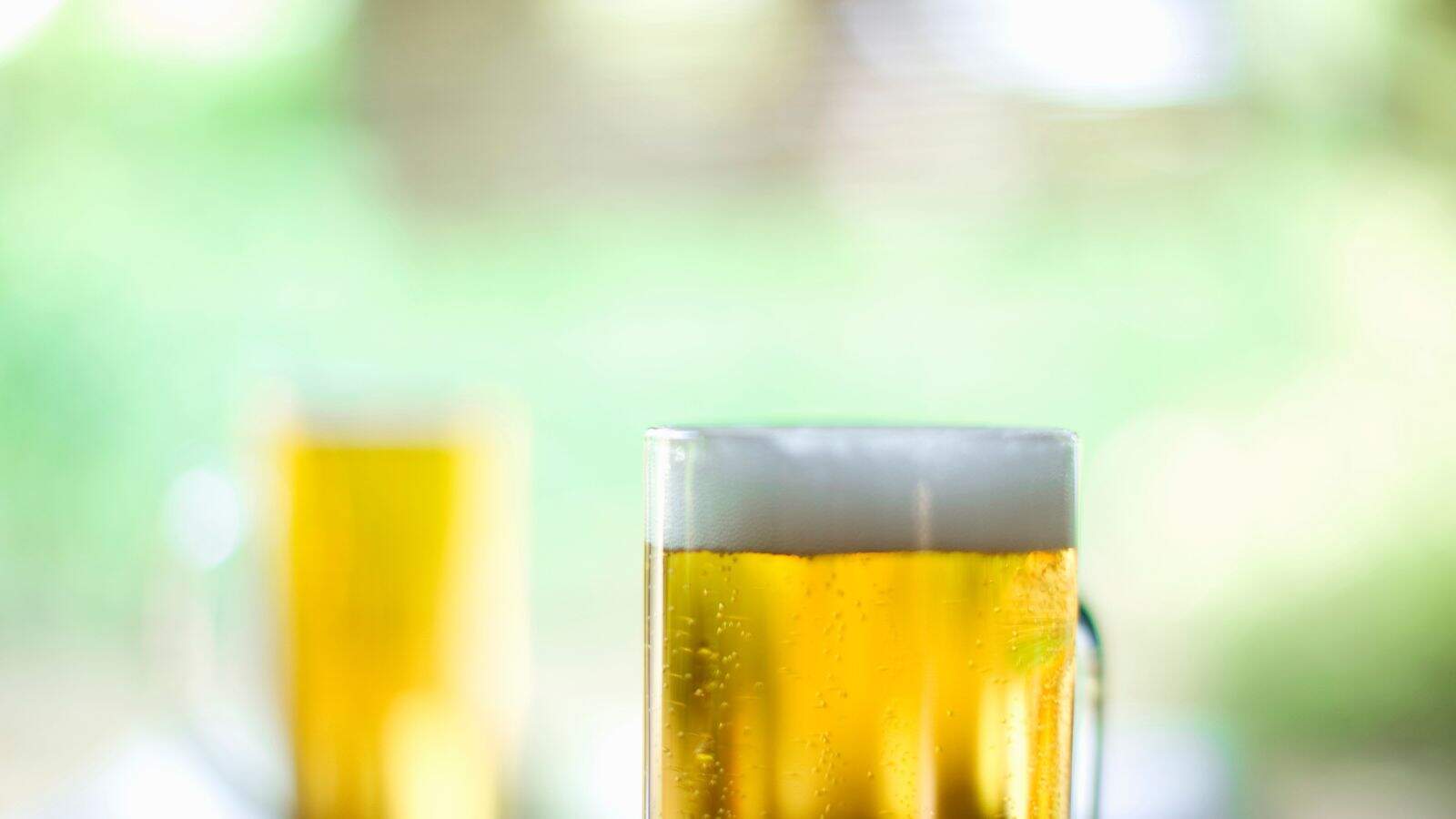 A glass mug filled with golden beer topped with a layer of foam, set against a blurred green background. Another similar mug is visible in the background, also out of focus.
