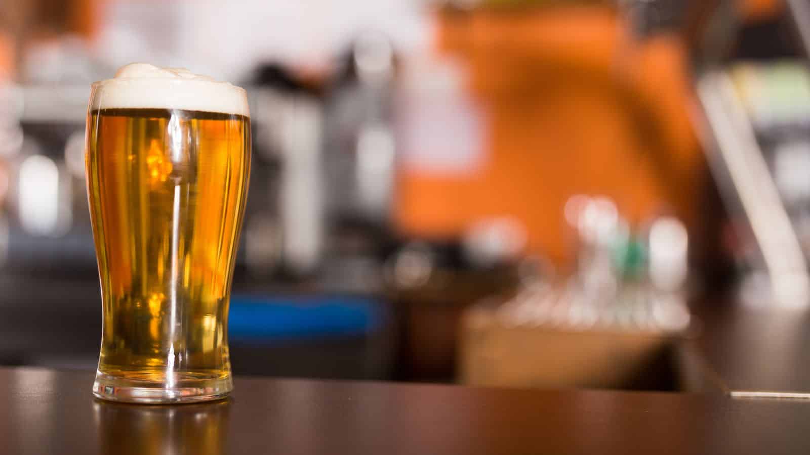 A clear glass of beer with a frothy head rests on a wooden bar counter. The background is blurred, featuring indistinct shapes and warm lighting, suggesting a bar or pub setting.