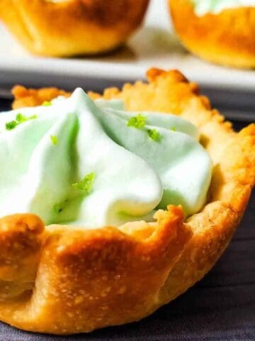 Close-up of a pastry tart filled with light green whipped cream and garnished with small green flakes. Several similar tarts are blurred in the background on a white tray, all set on a dark wooden surface.