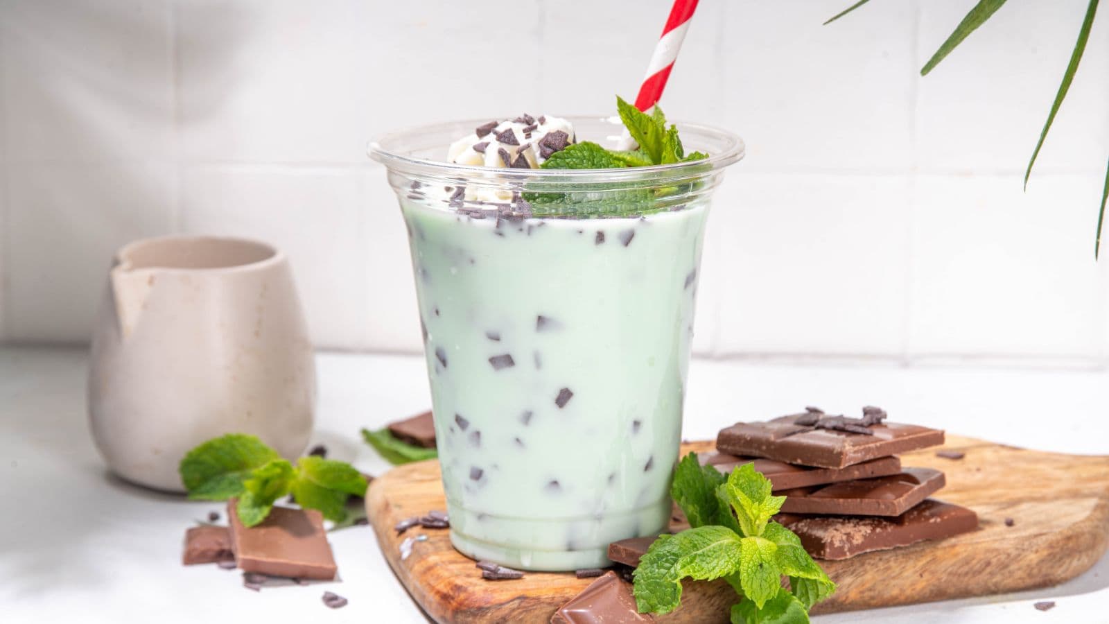 A green mint chocolate chip milkshake in a clear cup with a red and white striped straw, topped with whipped cream, mint leaves, and chocolate chips. Next to it, there are chocolate pieces and more mint leaves on a wooden board.