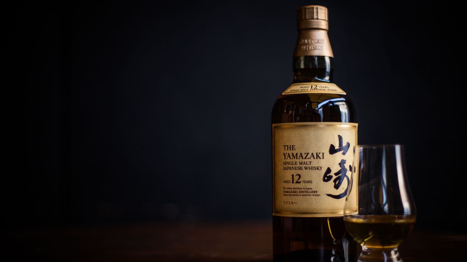 A bottle of The Yamazaki 12 Year Old Single Malt Japanese Whisky stands on a wooden surface next to a glass containing whisky, set against a dark background.