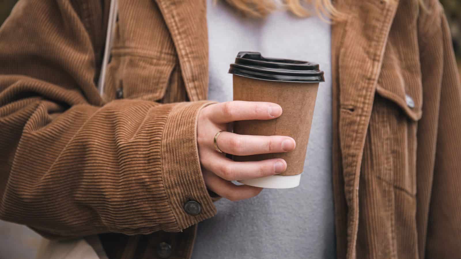A person wearing a brown corduroy jacket holds a brown paper coffee cup with a black plastic lid. A small part of a gray shirt is visible underneath the jacket. The person's hand has a gold ring on the index finger.