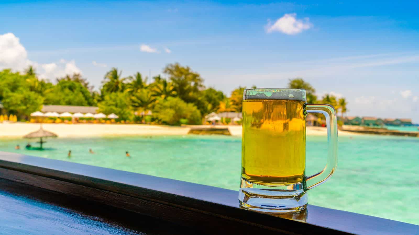 A glass mug of beer sits on a wooden railing overlooking a tropical beach with turquoise water, people swimming, and palm trees under a blue sky.